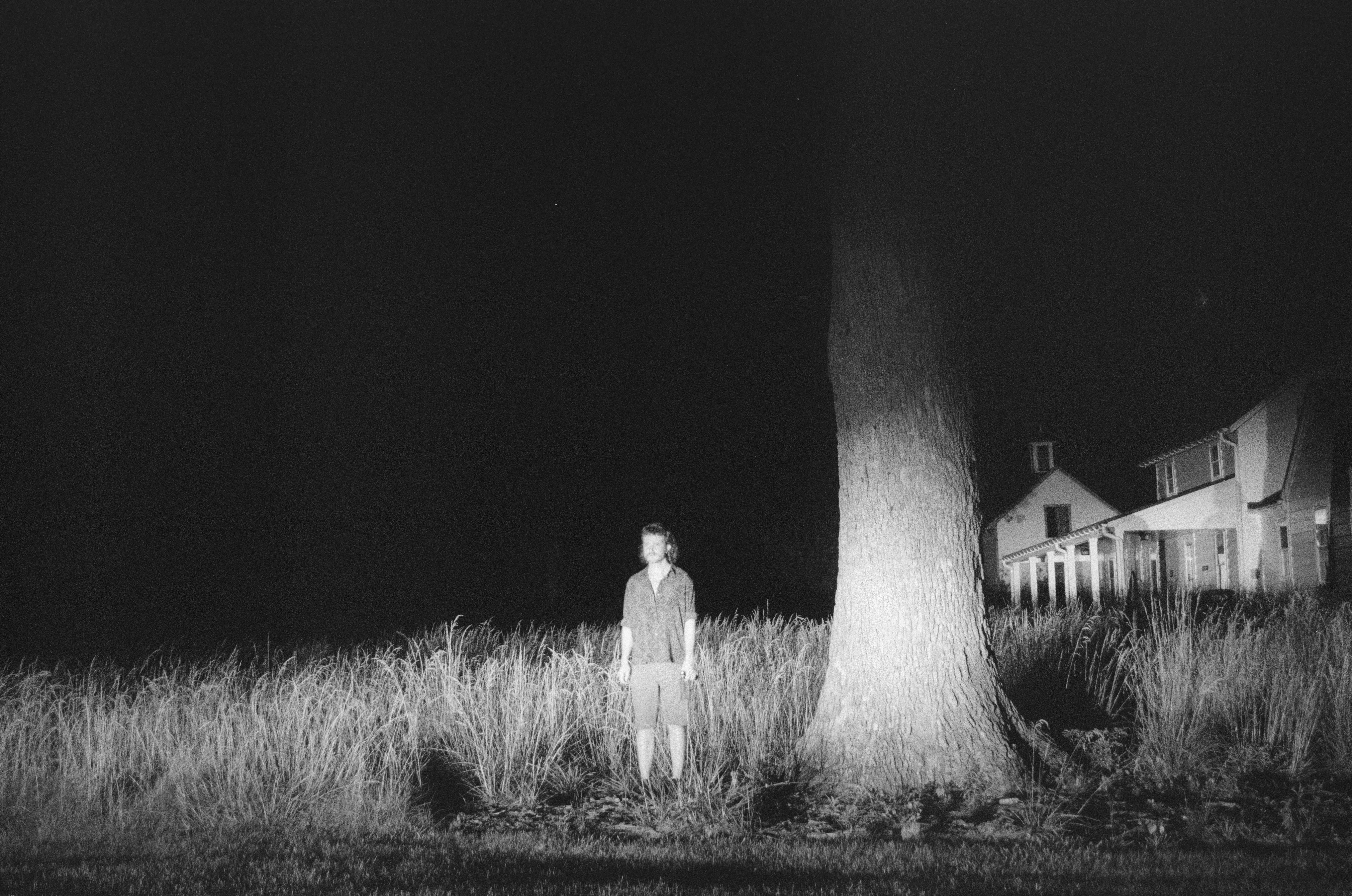 Ethan Evans standing next to the trunk of a large tree, at night, the scene illuminated by headlights with the stalks of grasses and an old looking home and barn visible in the background